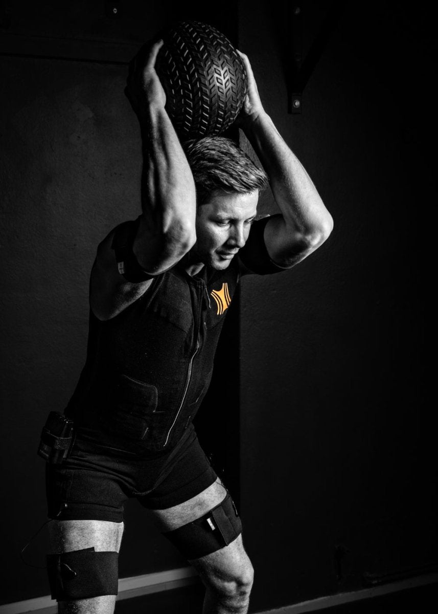 Ein Mann trägt den Kraftwunder EMA-Anzug und befindet sich in der Wurfbewegung mit einem Gewichtsball – Teil eines dynamischen Trainings im Rahmen von Therapiegold zur Aktivierung der Muskulatur.